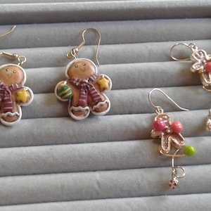May include: Three pairs of gingerbread man earrings. One pair features a gingerbread man with a red and white striped scarf and green and yellow ornaments. Another pair features a gingerbread man with a red and white striped scarf and green and yellow ornaments. The third pair features a gingerbread man with pink and white ornaments.