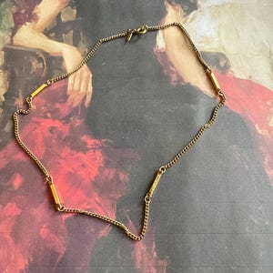 Puede incluir: Un collar de cadena dorada con cuatro colgantes rectangulares en forma de barra. El collar está sobre un fondo rojo y negro.