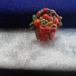 May include: A small, brown wicker basket filled with red and green fruit, with a red bow on the handle. The basket is made of plastic and has a realistic look.