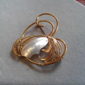 May include: A gold-toned brooch with a white, pearlescent leaf design. The brooch is in the shape of a heart with intricate filigree details.