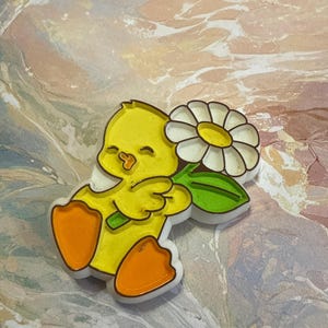 Puede incluir: Un pollito amarillo con patas naranjas sosteniendo una margarita blanca con un tallo verde. El pollito tiene una sonrisa con los ojos cerrados y está hecho de plástico.