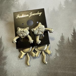 May include: A pair of silver cat earrings with a playful design. The cats are depicted in a jumping pose with their tails curled up. The earrings are on a black card with the text "Fashion Jewelry".