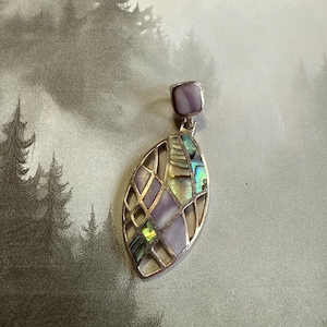 Può includere: Un ciondolo d'argento con un design geometrico con intarsi di madreperla di abalone iridescente. Il ciondolo ha la forma di una foglia e ha una conchiglia di abalone viola quadrata nella parte superiore.