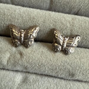 Puede incluir: Dos pendientes plateados con forma de mariposa. Cada pendiente presenta patrones detallados en las alas y un pequeño cuerpo. Se exhiben sobre una superficie gris texturizada, sugiriendo una exhibición o almacenamiento de joyas.