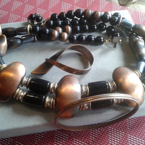 May include: A copper and black beaded necklace with a copper bracelet and a copper brooch. The necklace features large copper beads and black beads with gold accents.
