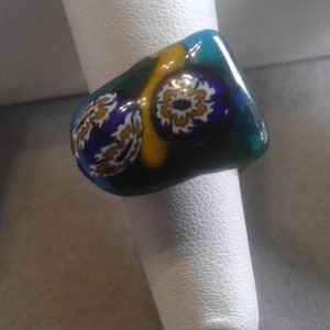 Könnte beinhalten: Ein Ring mit einer blauen, grünen, gelben und weißen Glasfassung. Die Fassung hat ein florales Design.