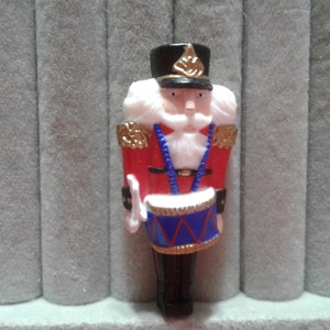 May include: A red and blue nutcracker figurine with a white beard and a black hat. The nutcracker is holding a drum with a red, white, and blue design.