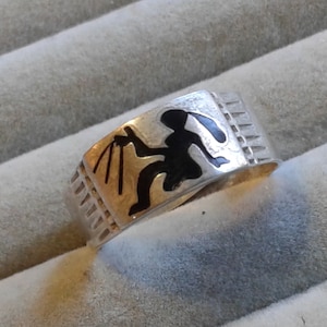 Puede incluir: Un anillo de plata con un diseño de esmalte negro de una persona sosteniendo un bastón. El anillo tiene una banda texturizada.