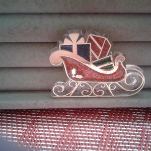 May include: A red and gold Christmas sleigh charm with a glittery finish. The sleigh is filled with colorful presents and has a white background.