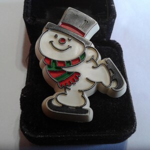Peut inclure: Une figurine de bonhomme de neige blanche portant un chapeau haut de forme noir, une écharpe rouge et verte et des patins à glace noirs. La figurine est dans une boîte en velours noir.