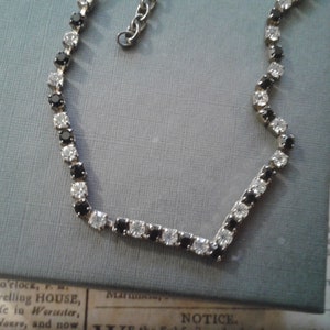 May include: A silver chain necklace with alternating black and white round rhinestones. The necklace is on a blue and white patterned surface.