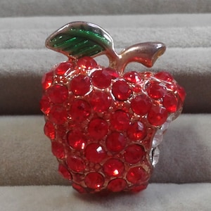 May include: A red apple pin with a green leaf, made of rhinestones. The apple is slightly bitten on one side.