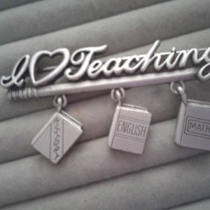 Puede incluir: Broche plateada con las palabras "I Love Teaching!" y tres dijes en forma de libros con las palabras "History", "English" y "Math".