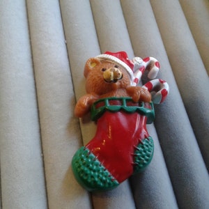 Könnte beinhalten: Ein brauner Teddybär mit einem rot-weißen Weihnachtsmann-Hut hält einen Zuckerstock und schaut aus einem roten Strumpf mit grünem Besatz.
