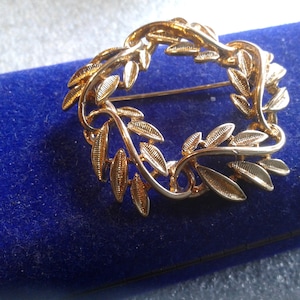 May include: A gold-toned brooch in the shape of a wreath of leaves. The brooch is on a blue velvet background.