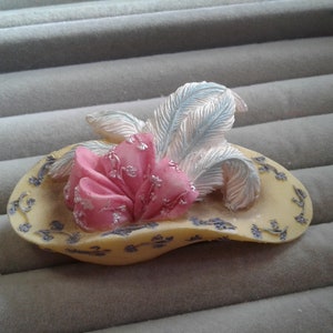 Puede incluir: Broche decorativo con forma de sombrero. El broche presenta una base amarillo pálido con detalles florales azules. Una flor de tela rosa y plumas plateadas adornan la parte superior. Un accesorio de inspiración vintage.