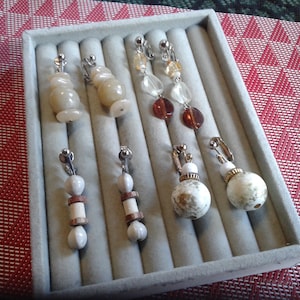 May include: A collection of clip-on earrings displayed in a gray jewelry tray. The earrings feature various designs, including stacked beads, dangling clear and amber-colored beads, and marbled spheres. The metal clips are silver-toned.