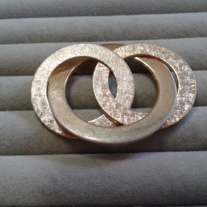 Può includere: Un fermaglio a forma di anello in metallo dorato con due cerchi intrecciati. I cerchi hanno una superficie testurizzata.