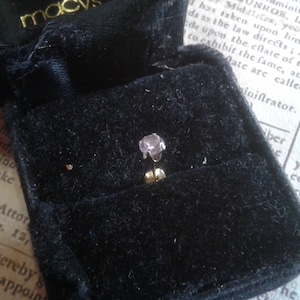 May include: A small, round, light purple gemstone earring in a black velvet jewelry box. The earring is set in a silver setting and has a post back.