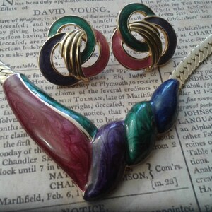 Puede incluir: Un collar de cadena de oro con un colgante multicolor. El colgante está hecho de esmalte y presenta un diseño de espiral en tonos de rojo, verde, morado y azul. El colgante está unido a la cadena con un cierre de oro.