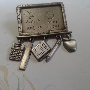 Puede incluir: Broche de metal plateado en forma de pizarra con ecuaciones y símbolos matemáticos. La pizarra tiene cinco colgantes: una calculadora, una regla, un libro con la palabra "MATH" en él, un lápiz y una manzana.