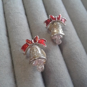 May include: A pair of silver-toned bell-shaped earrings with red enamel bows. The bells have a glittery texture and a small, clear gem at the bottom. The earrings are set against a gray, textured background.