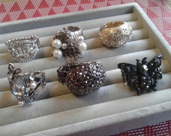 May include: Six silver rings with various designs, including a butterfly, a flower, and a geometric pattern. The rings are displayed on a gray velvet tray.