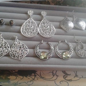May include: A collection of silver earrings with various designs, including filigree, geometric, and floral patterns. Some earrings feature clear crystals and green stones.