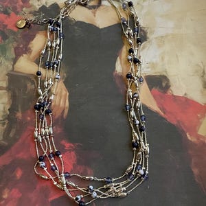 May include: A multi-strand necklace with silver-coloured metal beads and dark blue and white speckled beads. The necklace is set against a painting of a woman in a black dress.