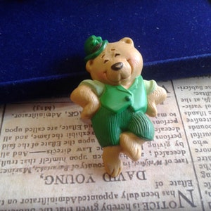 Könnte beinhalten: Eine grüne und braune Teddybär-Figur, die einen grünen Hut mit einem Kleeblatt auf der Oberseite trägt. Der Bär steht mit verschränkten Armen und trägt eine grüne Weste und eine grüne Hose.