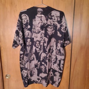 K&ouml;nnte beinhalten: Schwarzes T-Shirt mit einer Collage von Marilyn Monroe-Bildern in Grau- und Wei&szlig;t&ouml;nen. Die Bilder zeigen verschiedene Posen und bilden einen auff&auml;lligen Kontrast zum schwarzen Hintergrund. Das T-Shirt h&auml;ngt an einem Metallb&uuml;gel.