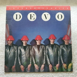 May include: A vinyl record album cover featuring the band DEVO. The cover art shows five men wearing red, cone-shaped hats and silver jackets. The album title is "Freedom of Choice".