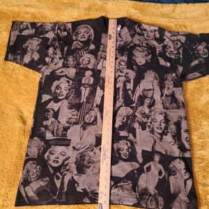 K&ouml;nnte beinhalten: Schwarzes T-Shirt mit einer Collage von Marilyn Monroe-Bildern in Grau- und Wei&szlig;t&ouml;nen. Das Design bedeckt das gesamte T-Shirt und erzeugt einen Vintage-Look. Das T-Shirt liegt flach auf einer gelben Oberfl&auml;che.