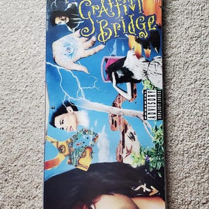 May include: A sealed VHS tape of Prince's "Graffiti Bridge" album. The cover features a collage of images with the album title in yellow script. The background is a mix of blue, orange, and green hues, with the Parental Advisory Explicit Lyrics label visible.