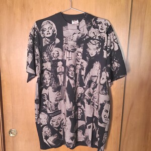 K&ouml;nnte beinhalten: Schwarzes T-Shirt mit einer Collage aus Schwarz-Wei&szlig;-Bildern von Marilyn Monroe. Die Bilder haben unterschiedliche Gr&ouml;&szlig;en und Posen und bilden ein einzigartiges Muster. Das T-Shirt h&auml;ngt an einem Metallb&uuml;gel.