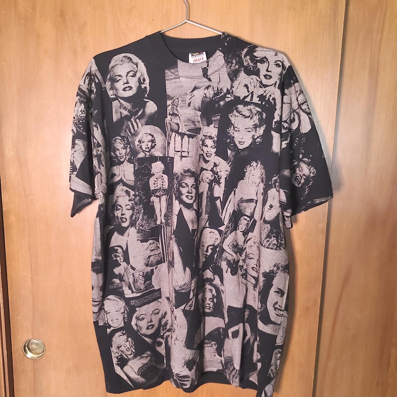 K&ouml;nnte beinhalten: Schwarzes T-Shirt mit einer Collage aus Schwarz-Wei&szlig;-Bildern von Marilyn Monroe. Die Bilder haben unterschiedliche Gr&ouml;&szlig;en und Posen und bilden ein einzigartiges Muster. Das T-Shirt h&auml;ngt an einem Metallb&uuml;gel.