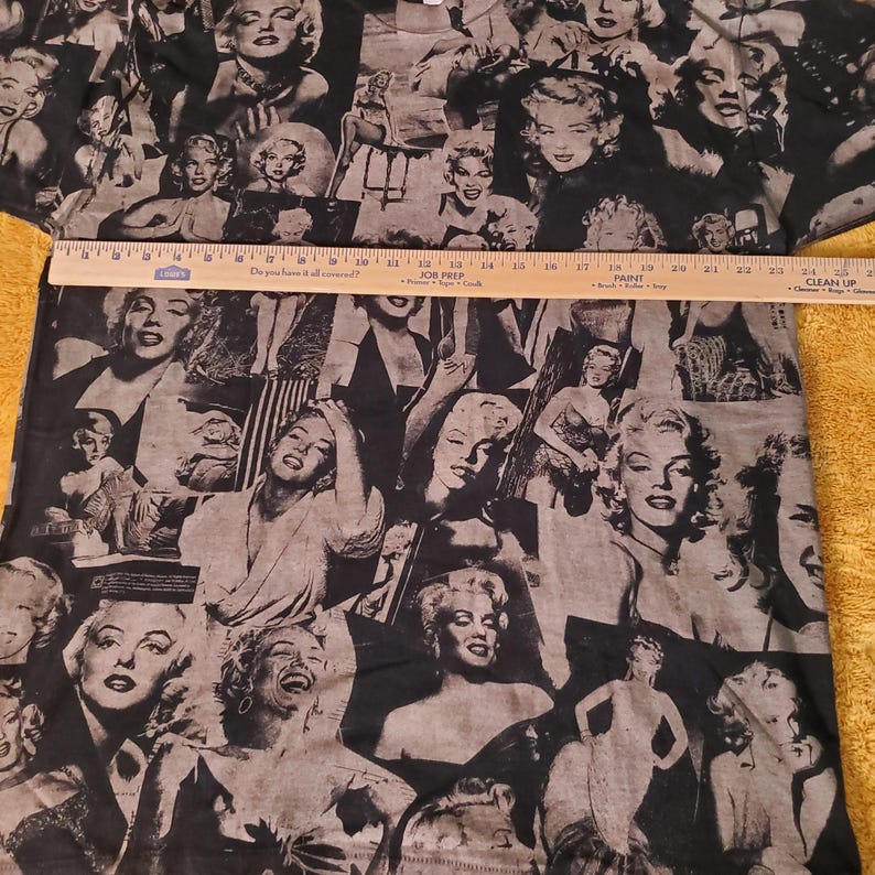 K&ouml;nnte beinhalten: Schwarz-graues T-Shirt mit einer Collage von Marilyn Monroe-Bildern. Das Design zeigt verschiedene Posen der Schauspielerin. Ein Lineal liegt quer &uuml;ber dem T-Shirt und misst etwa 61 cm.