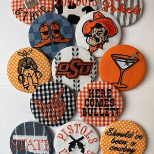 May include: A collection of thirteen circular fabric-covered buttons featuring embroidered designs related to Oklahoma State University, including text like 'Loyal & True', 'Cowboys', 'Go Pokes', 'OSU', 'Here Comes Bullet', 'State', 'Pistols Firing', and 'Should've been a cowboy'.
