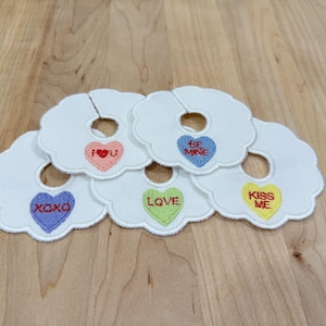 Peut inclure: Sous-verres blancs en forme de nuage avec des motifs de cœurs brodés. Chaque dessous de verre porte un message différent : "I U", "Be Mine", "XOXO", "Love" et "Kiss Me". Les dessous de verre sont disposés sur une surface en bois.