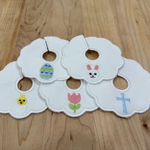 Peut inclure: Cinq bavoirs blancs à bord festonné, chacun orné de broderies sur le thème de Pâques. Les motifs comprennent un œuf de Pâques, un lapin, un poussin, une tulipe et une croix. Les bavoirs sont disposés sur une surface en bois.