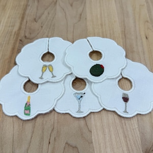 Peut inclure: Sous-verres blancs à bord festonné avec des motifs brodés. Chaque dessous de verre présente un cocktail différent : flûtes à champagne, une olive, un martini, un verre de vin et une bouteille de champagne. Les dessous de verre ont un trou au centre.