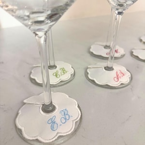 Peut inclure: Verres à martini transparents avec dessous de verre blancs à bords festonnés. Les dessous de verre comportent des initiales brodées en bleu, vert et rose. Les dessous de verre sont autour des verres.