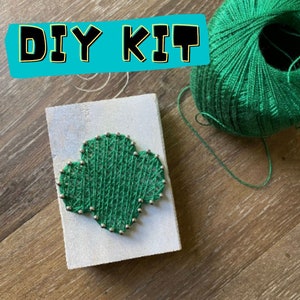 May include: DIY kit for making a string art project. The kit includes a wooden board, green yarn, and silver tacks. The yarn is wrapped around the tacks to create a green clover design.