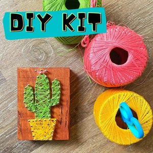 May include: DIY kit for string art featuring a green cactus with yellow pot on a wooden base. The kit includes three balls of yarn in pink, green, and yellow, and a pair of scissors.