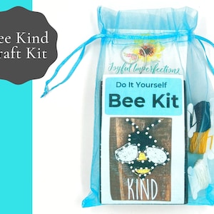 May include: A blue organza bag containing a craft kit to make a bee-themed sign. The kit includes a wooden sign with the word "KIND" printed on it, a pre-printed design of a bee, and embroidery floss in black, white, and yellow. The bag has a tag that reads "Joyful Imperfections Do It Yourself Bee Kit".