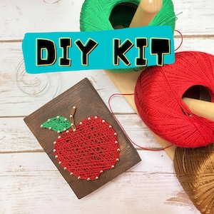 May include: DIY KIT text on a blue background with a wooden board with a red apple string art design. The apple is made with red yarn and has a green yarn leaf. There are three balls of yarn in the background: green, red, and brown.