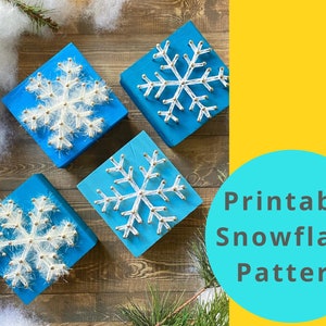 May include: Four blue square blocks with white snowflake designs made of string and small beads. The blocks are arranged on a wooden surface with greenery and white fluff. The image includes a teal circle with the text "Printable Snowflake Pattern."