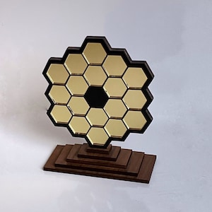 May include: A decorative model of the James Webb Space Telescope. The model features a gold-colored, hexagonal mirror array with a black central aperture, mounted on a tiered, brown wooden base. The telescope is a symbol of space exploration.