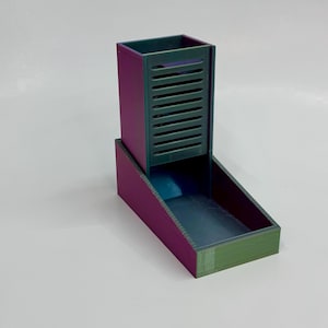 May include: A two-tone dice tower and tray. The tower is a dark teal with vertical slots, and the tray is a matching colour. The exterior is a gradient of purple and green. The dice tower is rectangular and sits atop a rectangular tray.
