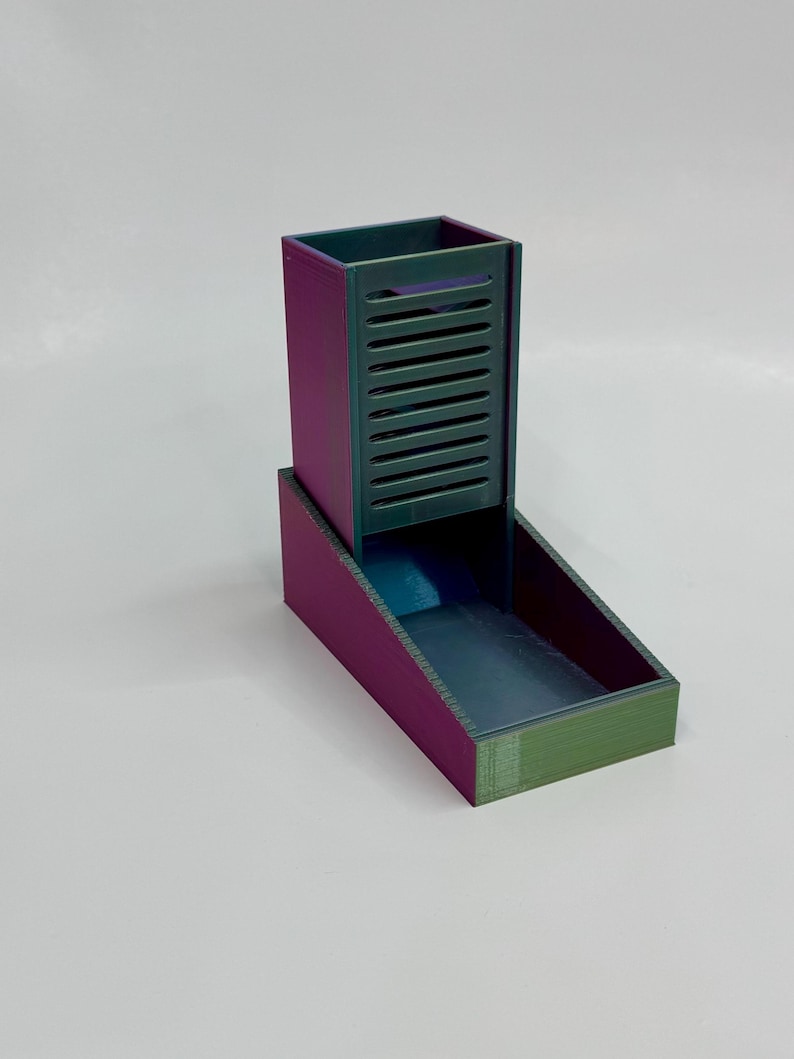 Op de afbeelding: Een 3D-geprinte dobbelsteentoren met een kleurveranderende afwerking die varieert tussen paars en blauwgroen. De structuur heeft een hoog, rechthoekig verticaal gedeelte met horizontale lamellen aan de voorzijde en een open bak aan de basis.